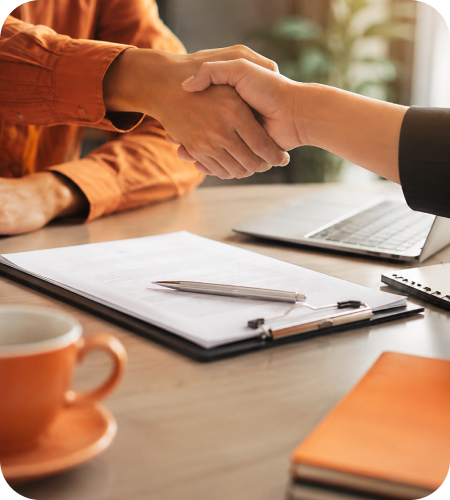 Two corporate professionals shaking hands over a wooden desk with a printed contract on a clipboard, laptop, executive pen, coffee mug, and orange notebook