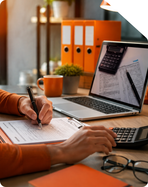 Tax professional reviewing financial records with a pen and calculator next to a laptop displaying income tax forms